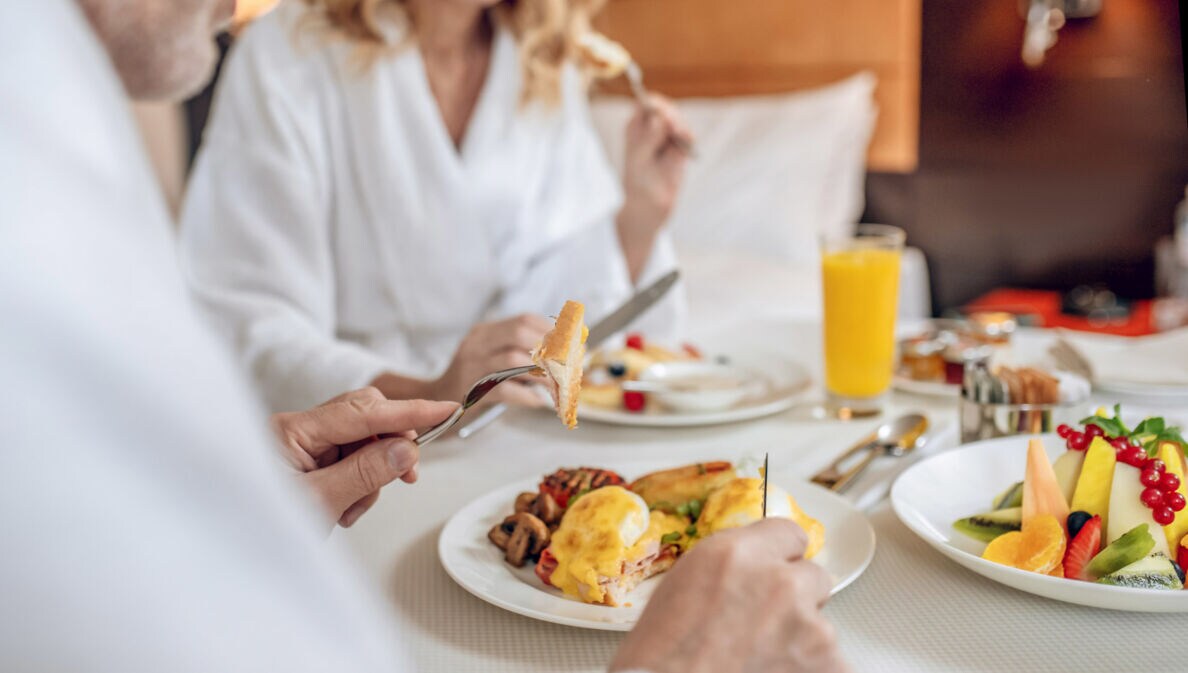 Nahaufnahme von zwei gesichtslosen Personen in weißen Bademänteln, die an einem eingedeckten Tisch in einem Hotelzimmer frühstücken.