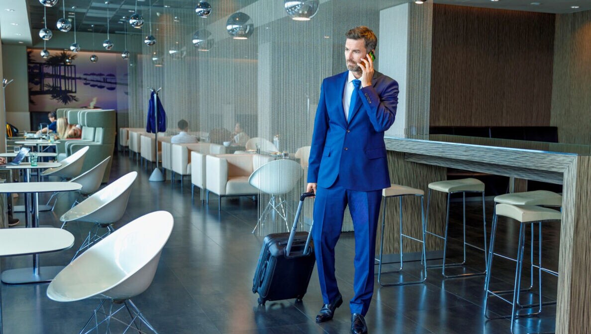 Ein Geschäftsmann im blauen Anzug und mit Trolley telefoniert stehend in einer modernen Flughafen-Lounge.