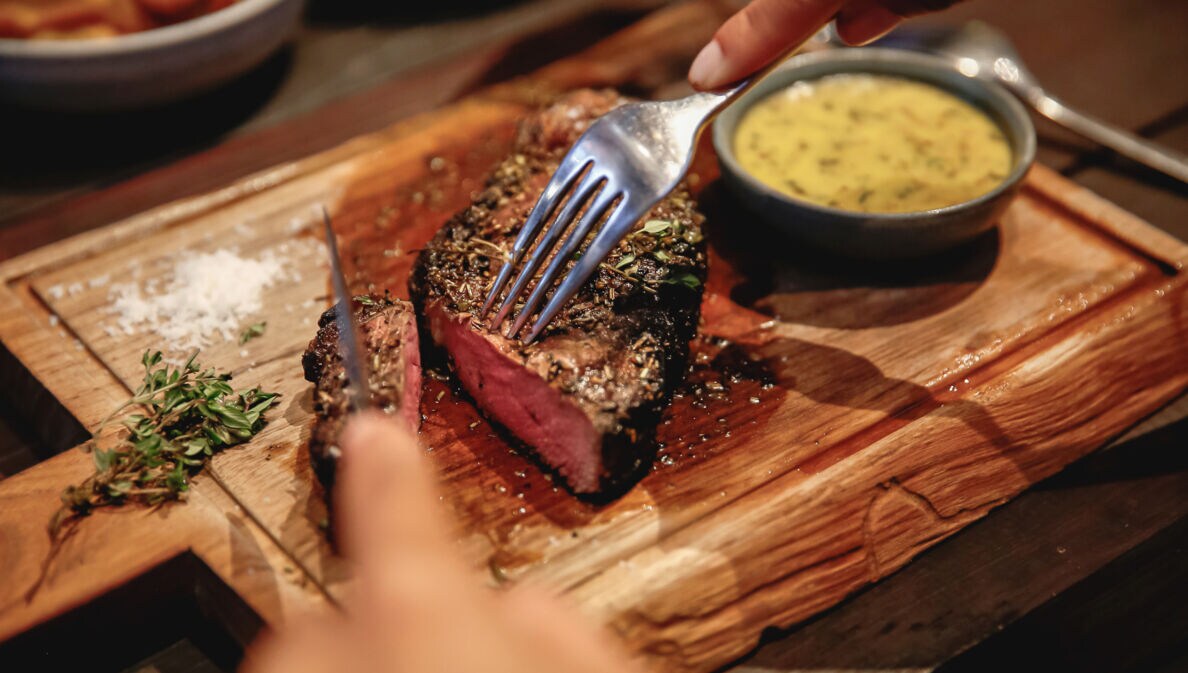 Nahaufnahme eines angeschnittenen Steaks auf einem Holzbrett neben einer Schale mit Soße.