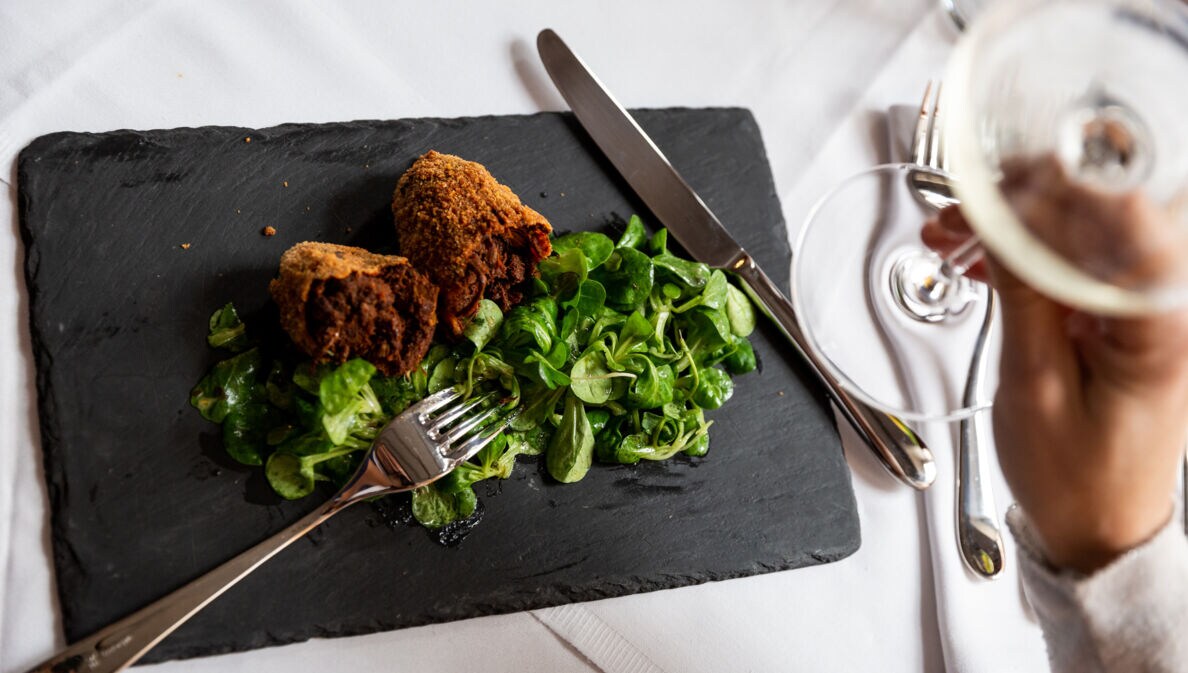 Aufsicht eines panierten Fleischgerichtes an Feldsalat auf einer Schieferplatte auf einem edel gedeckten Tisch, im Vordergrund eine Hand mit einem Glas Weißwein.