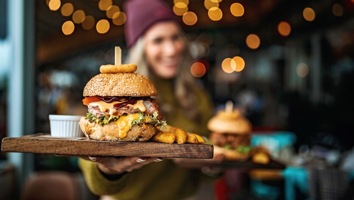 Ein Burger auf einem Brett, gehalten von einer Frau in Unschärfe in einem Lokal.