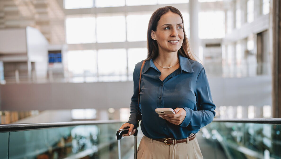 Eine lächelnde, elegant gekleidete Frau mit Smartphone und Trolley auf einer Rolltreppe in einem Flughafenterminal.