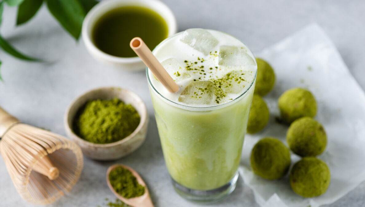 Ein Glas mit Matcha Latte und Strohhalm, daneben liegen grüne Kugeln, eine Schale mit grünem Pulver, ein Bambusbesen und eine Schale mit purem Matchatee.