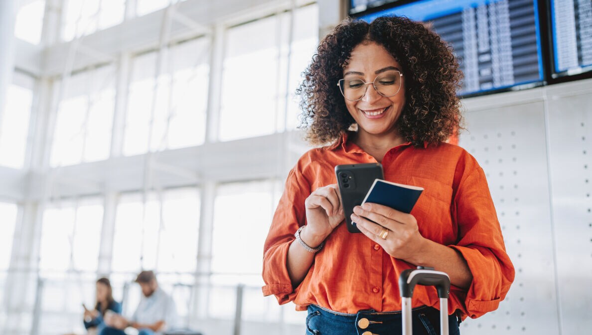 ine Frau bedient lächelnd ihr Smartphone in einem Flughafenterminal, in ihrer Hand ein Reisepass.