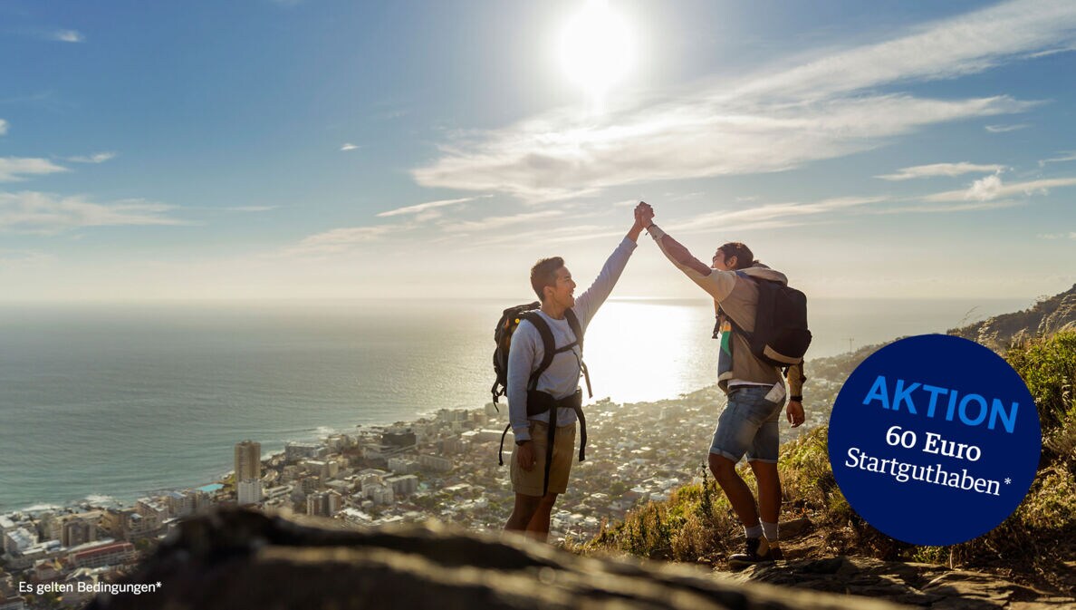 Zwei Wanderer mit Rucksäcken geben sich ein High Five auf einer Bergspitze, im Hintergrund eine Großstadt am Meer.