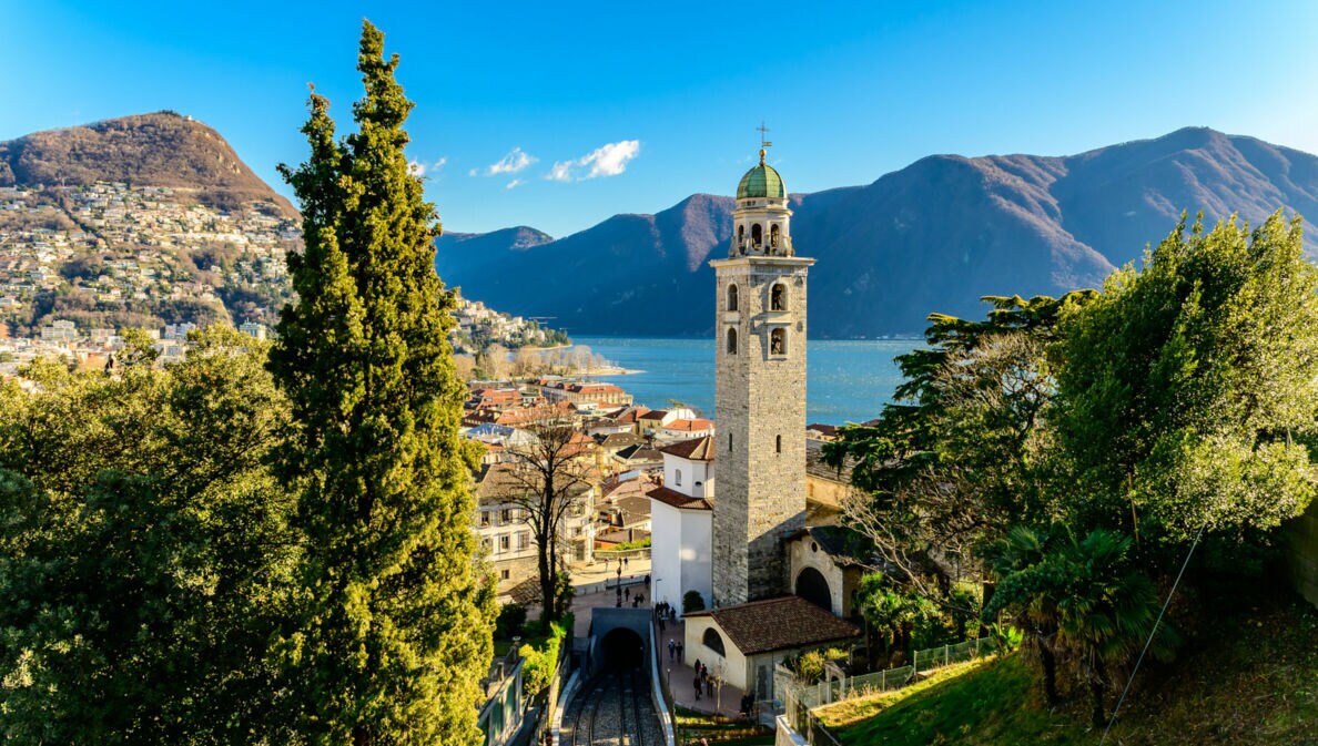 Stadtpanorama von Merano mit Zypressen und Glockenturm an einem See vor Bergpanorama.