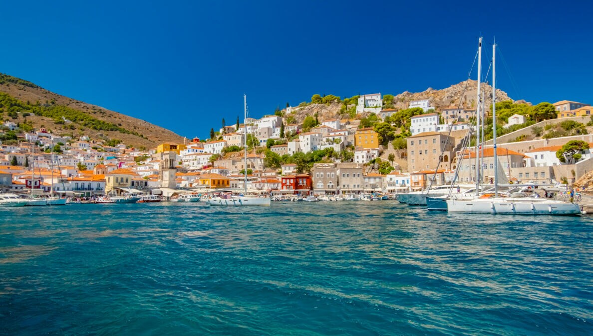 Mediterrane Ortschaft in Hügellage mit Hafenpromenade, im Vordergrund türkisblaues Meer mit Booten.