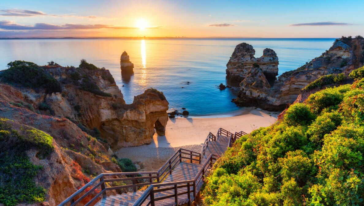 Der Strand Praia do Camilo in Portugal bei Sonnenuntergang, zu dem über Felsen eine Holztreppe nach unten führt.