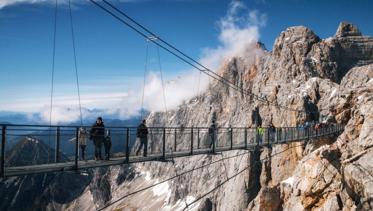 Personen auf einer Hängebrücke vor einer Felswand im Gebirge.