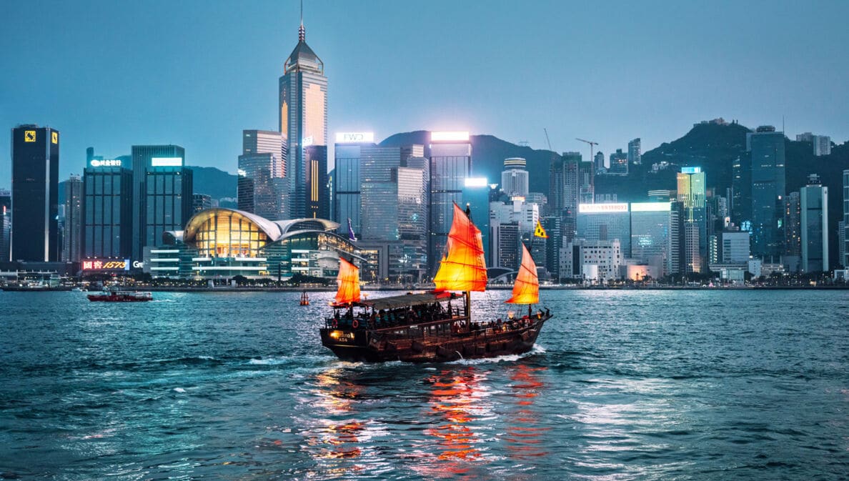 Rote chinesische Dschunke auf dem Wasser vor der Skyline Hongkongs bei Nacht.