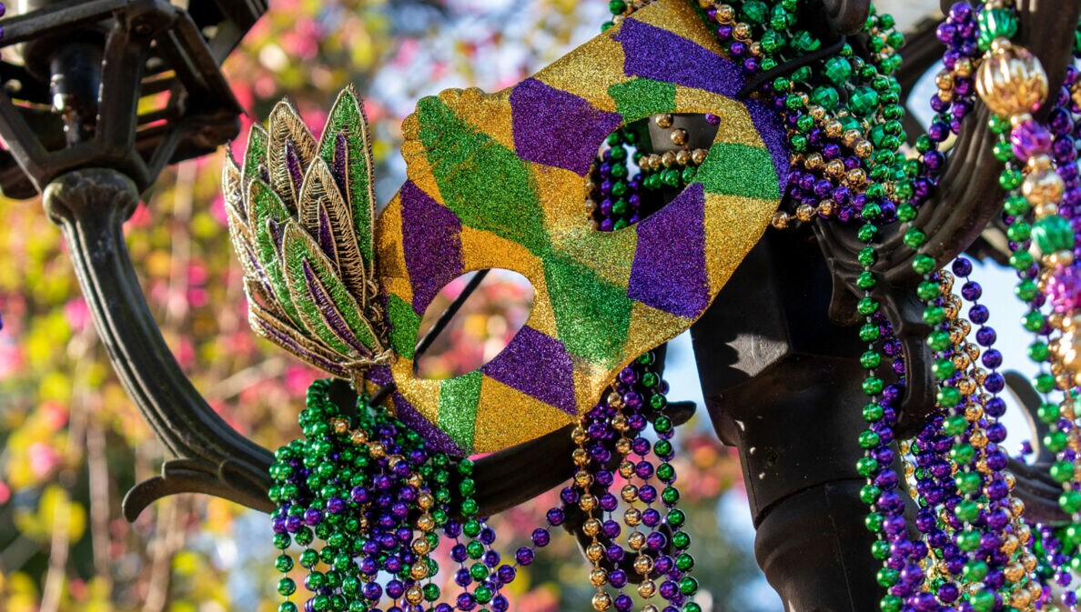 Eine Maske und Perlenketten in den Farben Grün, Gold und Violett an einer Straßenlaterne.