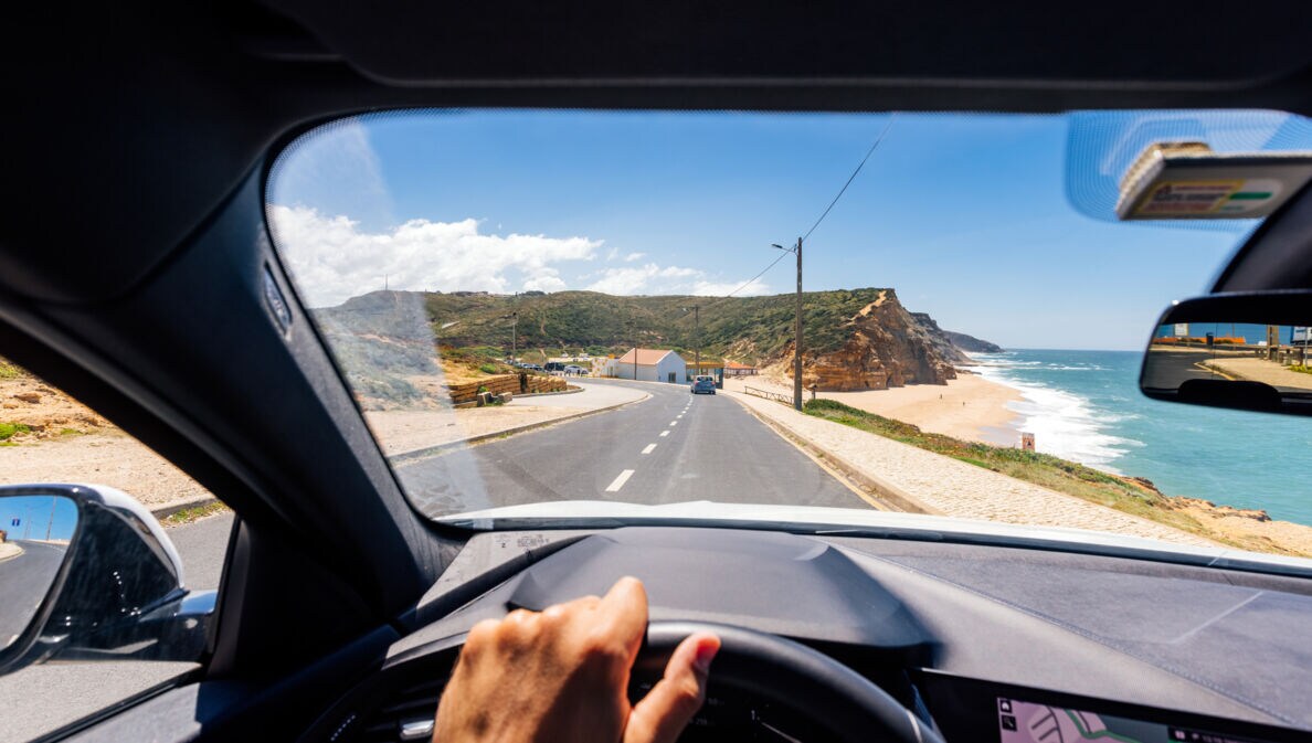 Blick über ein Lenkrad durch die Frontscheibe eines fahrenden Autos auf einer Straße an einer Küste mit Sandstrand bei Sonnenschein.