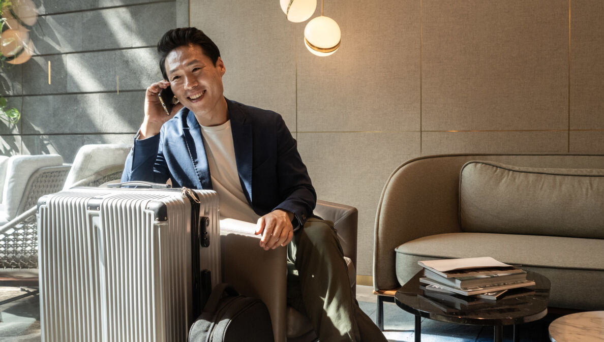 Ein fröhlicher asiatischer Geschäftsmann sitzt im Sessel einer modernen Flughafen-Lounge und telefoniert an seinem Smartphone, neben ihm steht ein großer Reisekoffer.