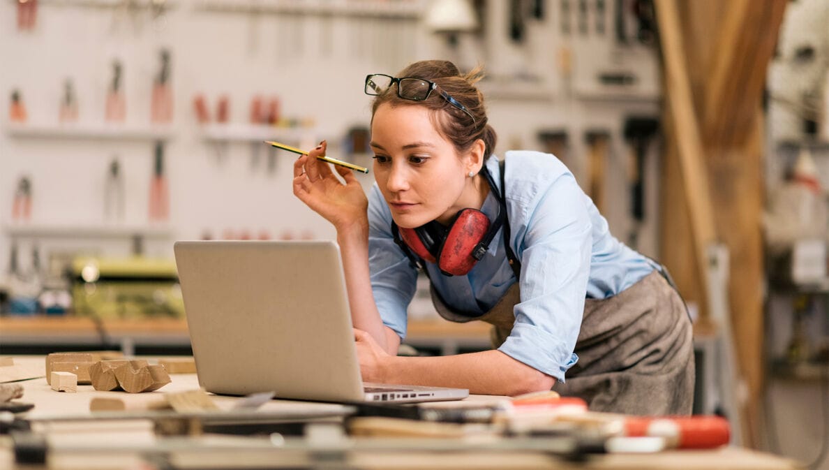 Foto einer Handwerkerin, wie sie an ihrer Werkbank steht und auf den vor ihr geöffneten Laptop blickt.