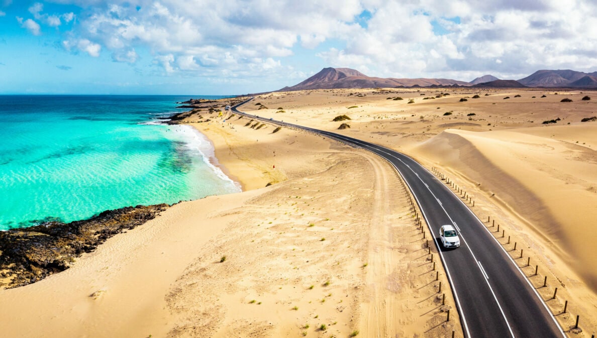 Ein weißes Auto fährt auf einer Schnellstraße durch eine Wüstenlandschaft am türkisblauen Meer.