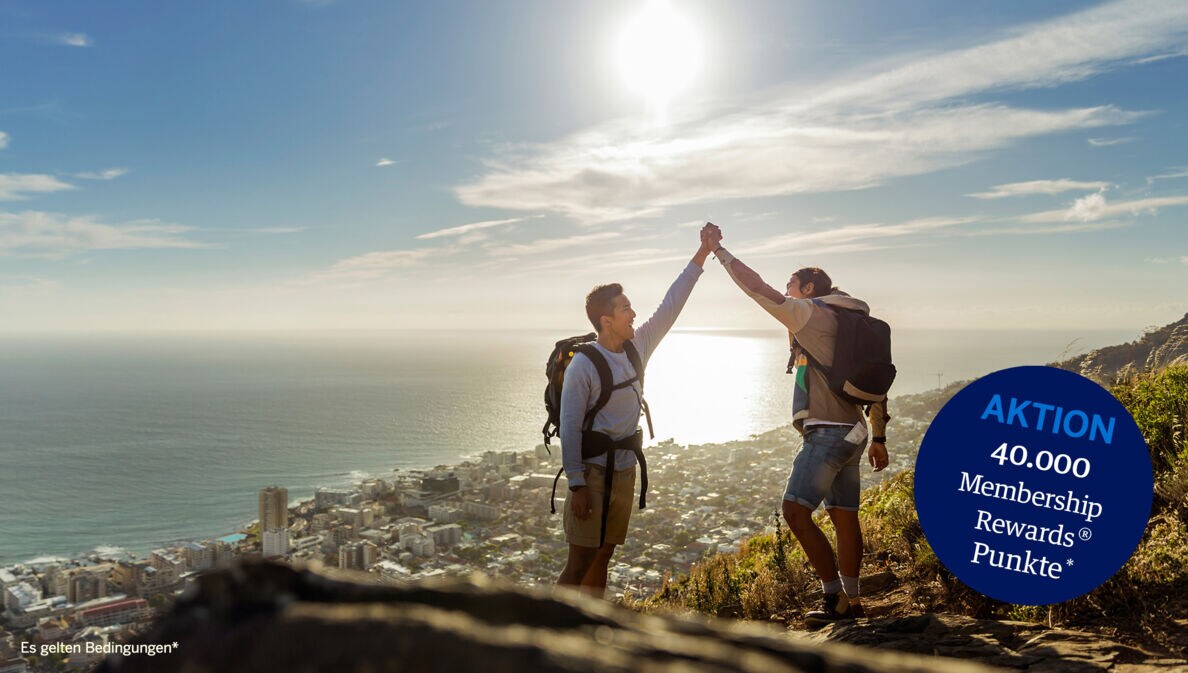 Zwei Wanderer mit Rucksäcken geben sich ein High Five auf einer Bergspitze, im Hintergrund eine Großstadt am Meer.