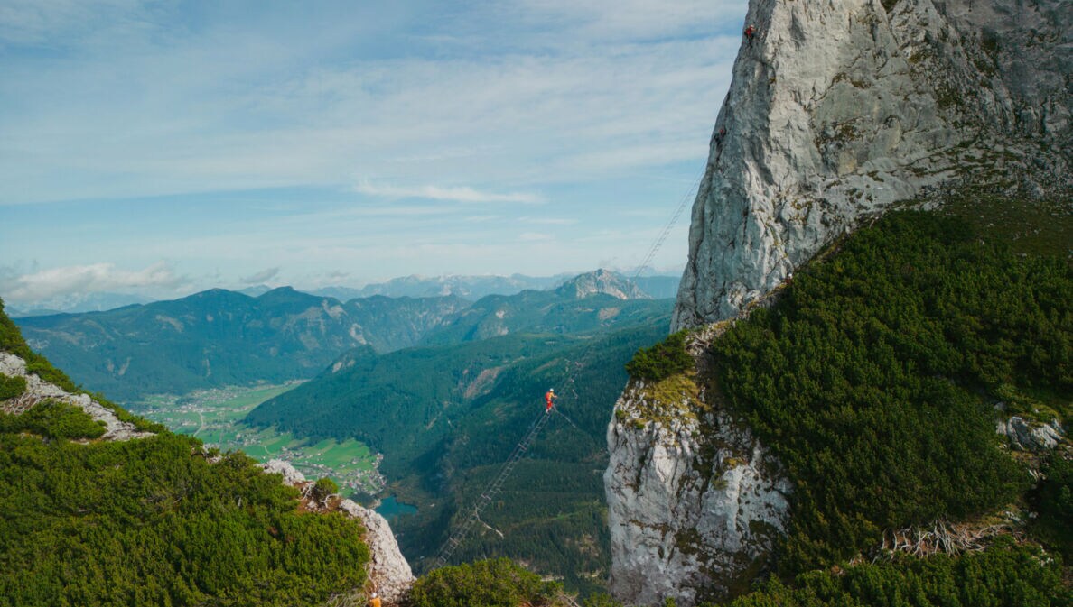 Eine Person auf einer Leiter, die hoch über einem Abgrund zu einem hohen Felsen führt, im Hintergrund Berge und Himmel.