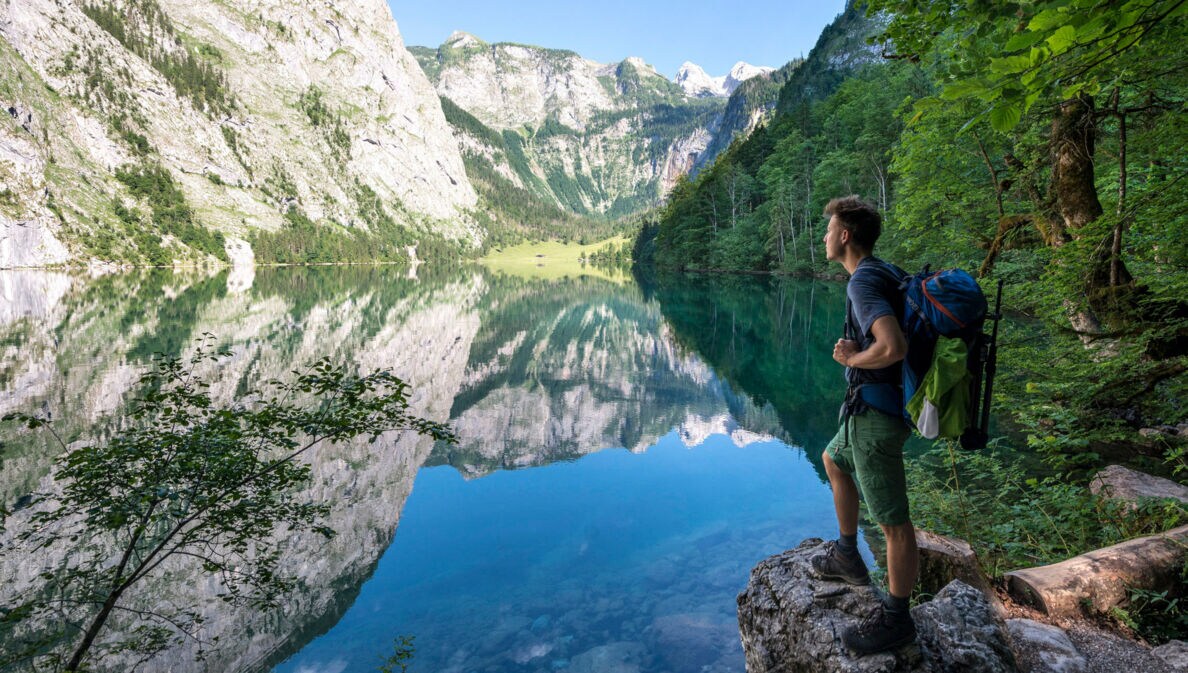 Eine Person mit Wanderrucksack steht auf einem Stein vor dem Königssee mit Blick auf die Berge.