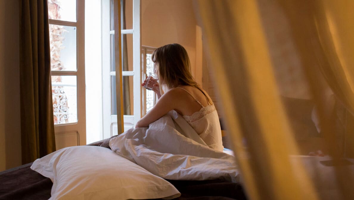 Rückenansicht einer Frau mit Wasserglas, die auf einem Bett sitzt und aus dem geöffneten Fenster schaut.