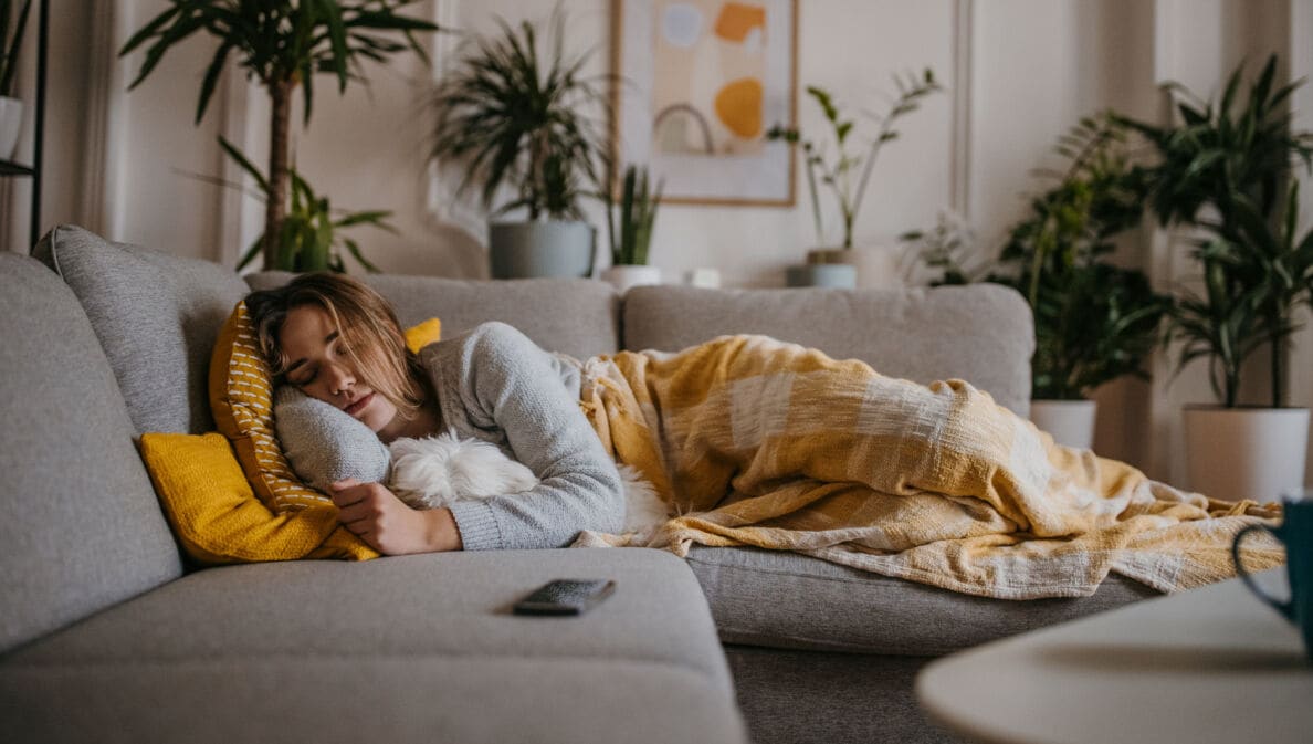 Eine Frau liegt schlafend mit einer Decke auf einer grauen Couch, im Arm hält sie einen Hund.