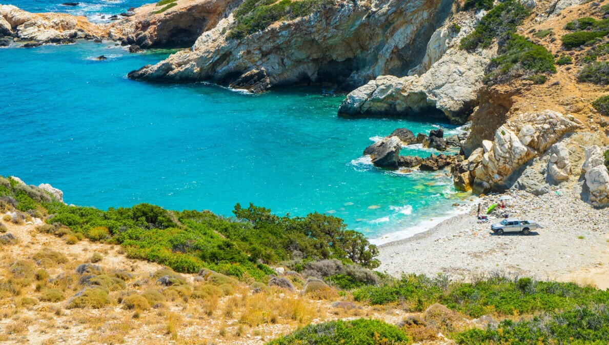 Ein Van steht auf einem Sandstrand in einer kleinen mediterranen Felsbucht mit türkisblauem Wasser.