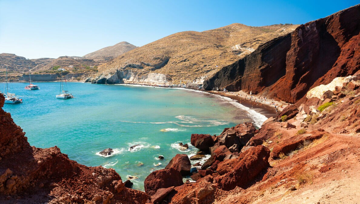 Meeresbucht mit Booten im türkisblauen Wasser, umgeben von roten Felsklippen an rotem, belebtem Sandstrand.