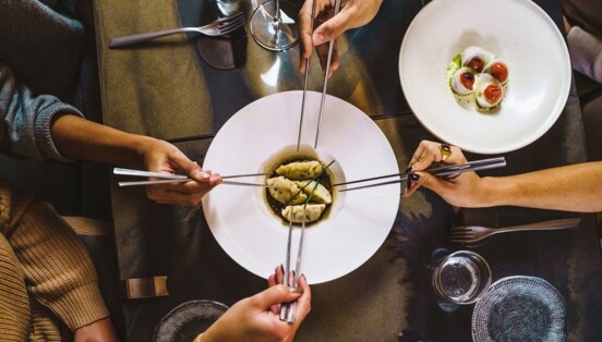 Aufsicht eines gedeckten Restauranttisches mit einem weißen Teller mit Teigtaschen, umgeben von vier Händen mit Essstäbchen.