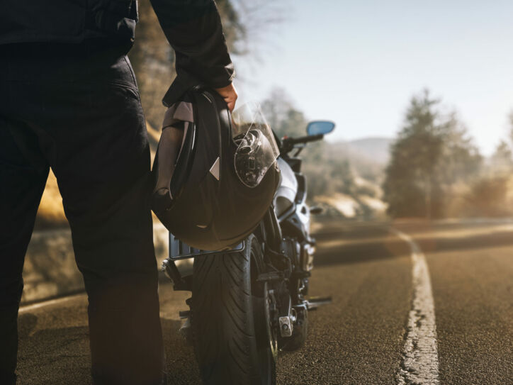 Ein Mann mit Motorradhelm in der Hand steht auf einer einsamen Landstraße vor einem Motorrad.