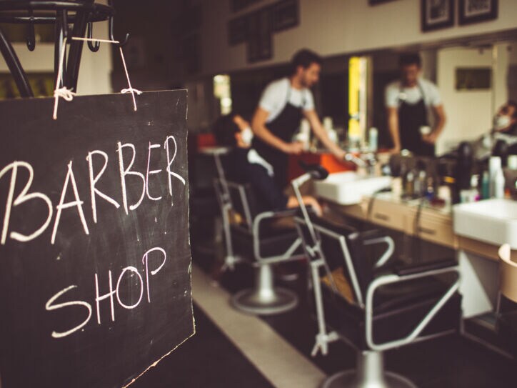 Eine Tafel auf der mit Kreide Barbershop steht, im Hintergrund ist ein Barbier mit seinem Kunden im Laden zu sehen