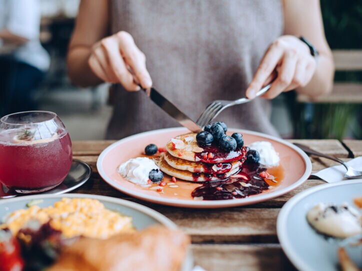 Eine Person sitzt an einem gedeckten Tisch vor einem Teller mit Pancakes im Außenbereich eines Cafés