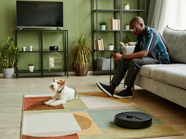 Ein Mann sitzt im Wohnzimmer auf dem Sofa und schaut auf sein Handy in der Hand. Vor ihm liegt ein Hund auf dem Teppich, daneben arbeitet ein Saugroboter.