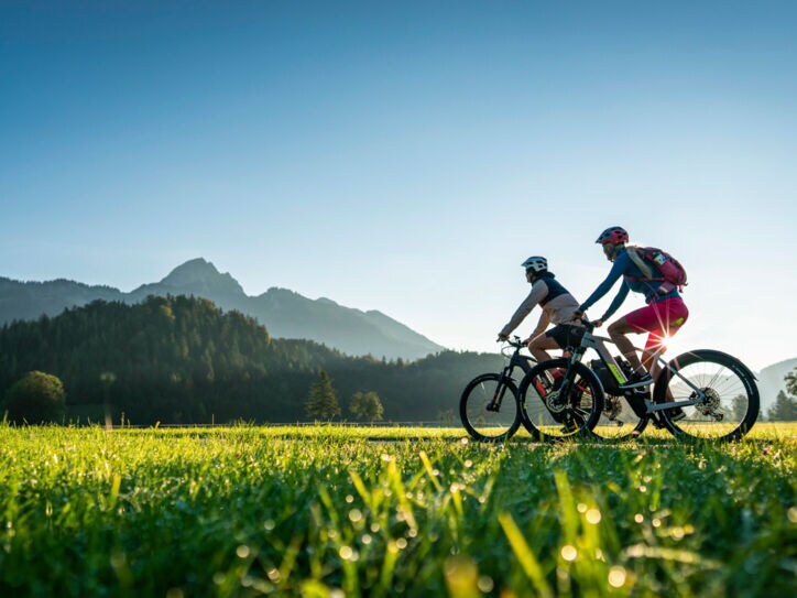 Zwei Personen fahren auf Mountainbikes durch eine Grasebene vor Bergpanorama im Sonnenschein