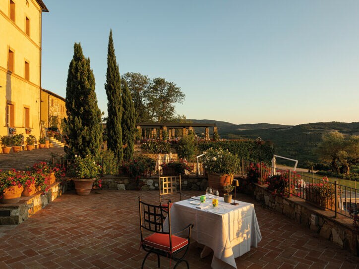 Ein gedeckter Tisch auf einer großen Terrasse einer Villa im toskanischen Baustil mit Blick in eine grüne Hügellandschaft bei Sonnenuntergang