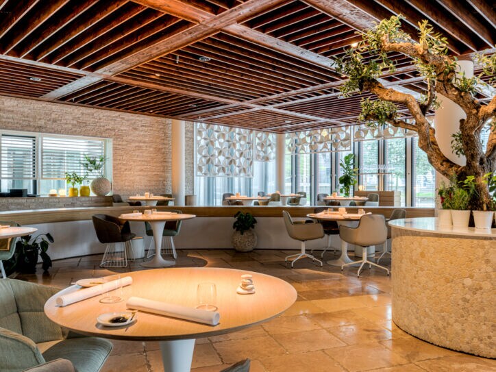 Modernes Restaurant mit runden Tischen und einem Olivenbaum in einem Rundbeet.