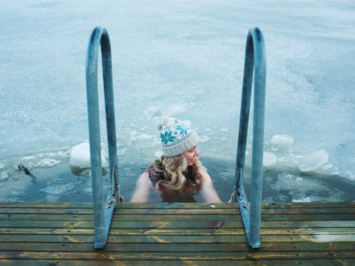 Eine Frau mit Wollmütze hält sich beim Baden in einem zugefrorenen See an dem Handlauf einer Treppe an einem Holzsteg fest.