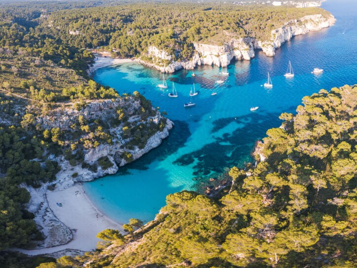 Luftaufnahme eines Küstenabschnitts von Menorca mit Badebuchten, Waldlandschaft und Booten.