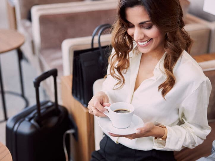 Eine elegant gekleidete Frau sitzt mit einer Tasse Kaffee in einem Sessel, neben ihr steht Gepäck.