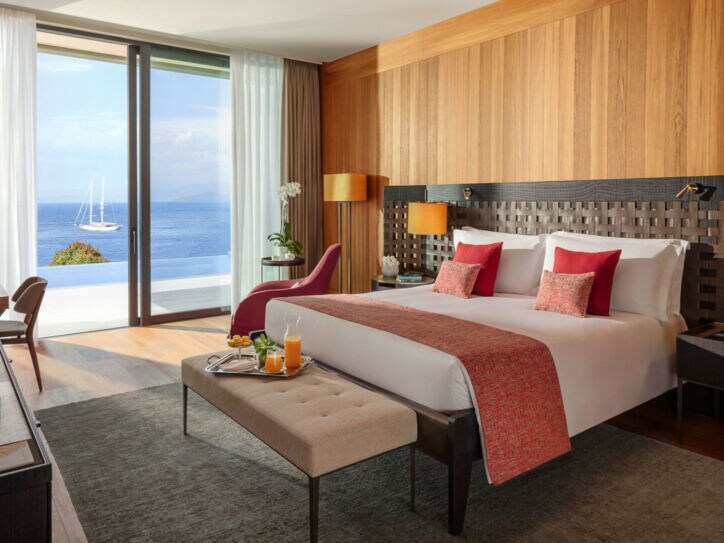 Modern eingerichtete Suite des Hotels Mandarin Oriental Bodrum mit einem großen Bett, einer Sitzbank, Arbeitstisch und raumhohen Fenstern mit Meeresblick.