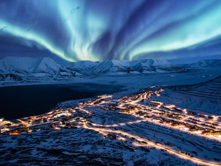 Beleuchtete Ortschaft in schneebedeckter Fjordlandschaft unter Polarlichtern bei Nacht.