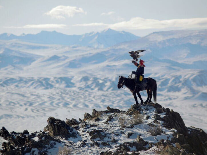 Ein Mann sitzt auf einem Pferd im Gebirge, auf seinem ausgestreckten Arm steht ein Adler.