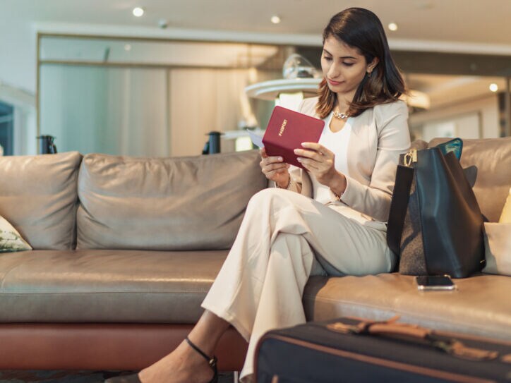 Eine Frau sitzt mit überschlagenen Beinen auf einem hellen Ledersofa und sieht sich Reiseunterlagen an.