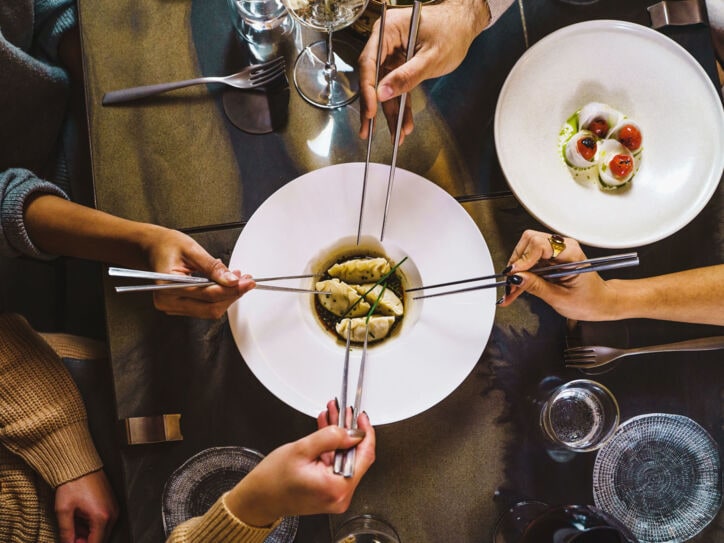 Aufsicht eines gedeckten Restauranttisches mit einem weißen Teller mit Teigtaschen, umgeben von vier Händen mit Essstäbchen.