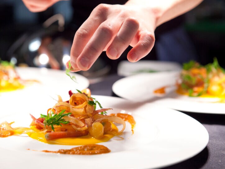 Eine Hand garniert ein Gericht auf einem weißen Teller in einer Restaurantküche.