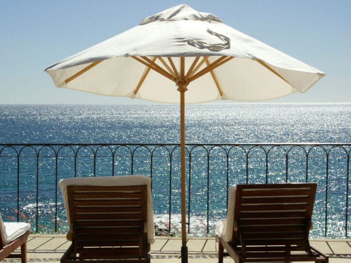 Terrasse und Geländer mit Blick aufs Meer, auf der mehrere Liegestühle und ein weißer Sonnenschirm stehen.