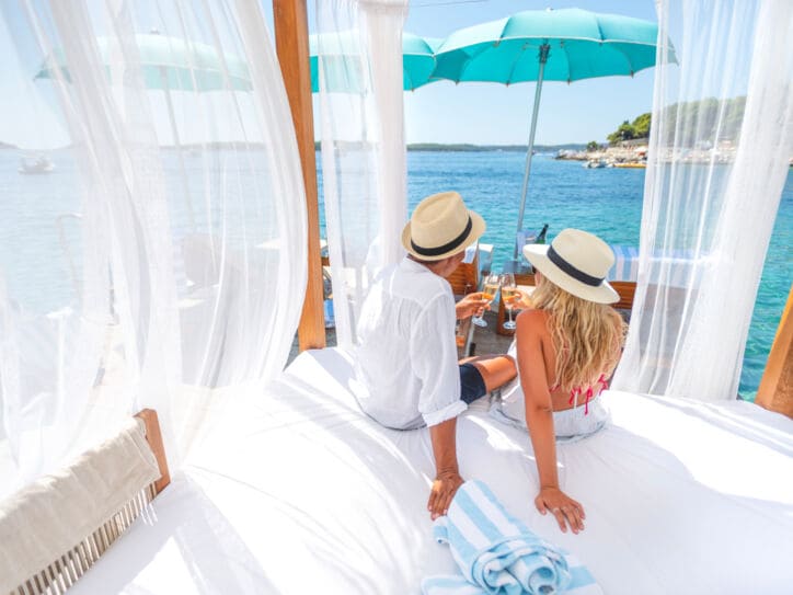 Rückansicht eines Paares mit Strohhüten und Champagnergläsern auf einem Daybed am Meer.