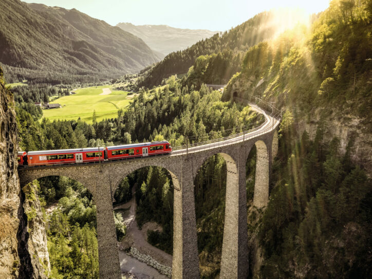 Ein roter Zug fährt auf einem Viadukt durch eine grüne Berglandschaft.