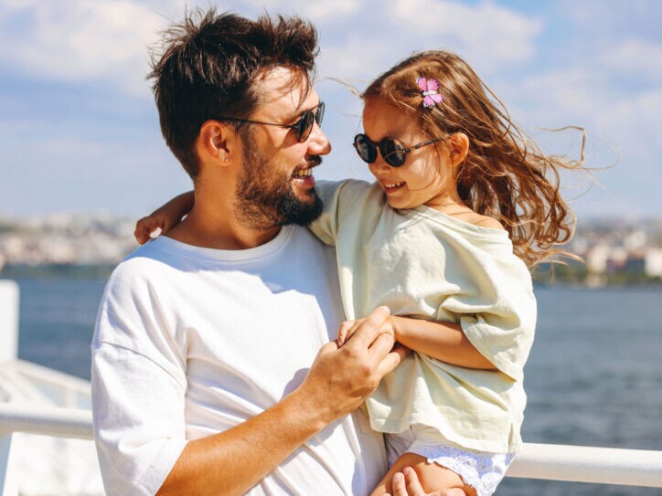 Ein lächelnder Mann mit Sonnbrille mit einem fröhlichen Mädchen mit Sonnenbrille auf dem Arm auf dem Außendeck eines Schiffes.