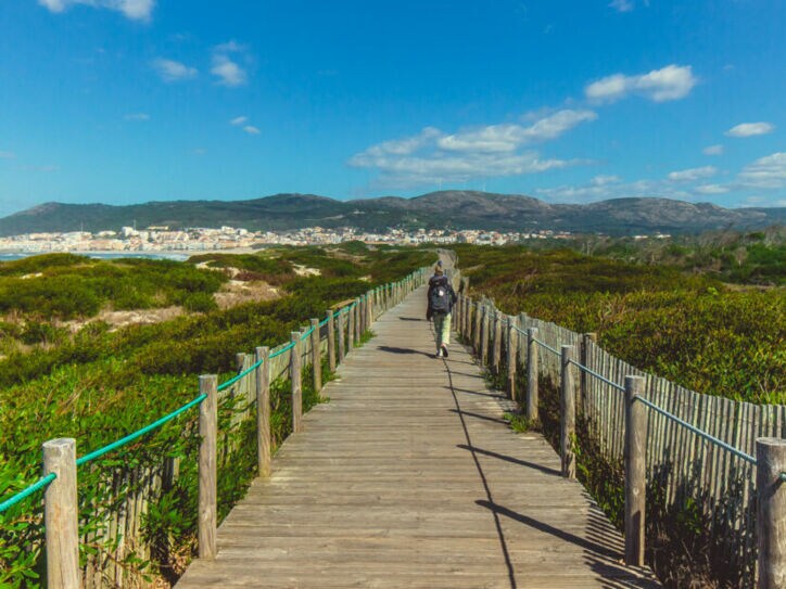 Eine unkenntliche Person beim Wandern in Portugal auf einem Holzsteg, der in Küstennähe über grüne Vegetation führt.