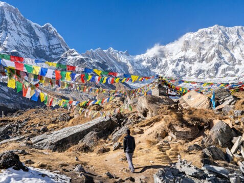 Rückansicht eines Mannes in einer schroffen Gebirgslandschaft, umgeben von bunten tibetischen Gebetsfahnen.