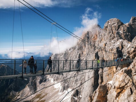 Personen auf einer Hängebrücke vor einer Felswand im Gebirge.